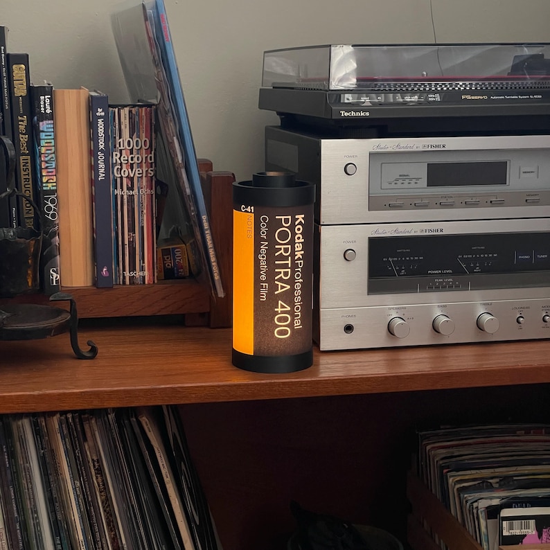 May include: A Kodak Professional PORTRA 400 color negative film canister stands on a wooden shelf, next to books and audio equipment. The canister is black with orange and white text. A record player and stereo components are in the background.