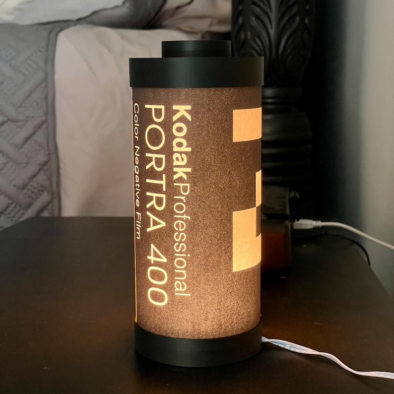 May include: A cylindrical lamp designed to resemble a Kodak Professional Portra 400 film canister. The lamp has a black base and top, with the film details printed on the brown body. The lamp is lit from within, creating a warm glow.