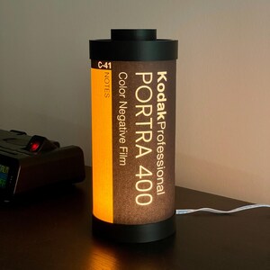 May include: A cylindrical lamp designed to resemble a Kodak Professional Portra 400 film canister. The lamp features a black top and base, with the film details printed on a brown and orange background. A white cord extends from the base.
