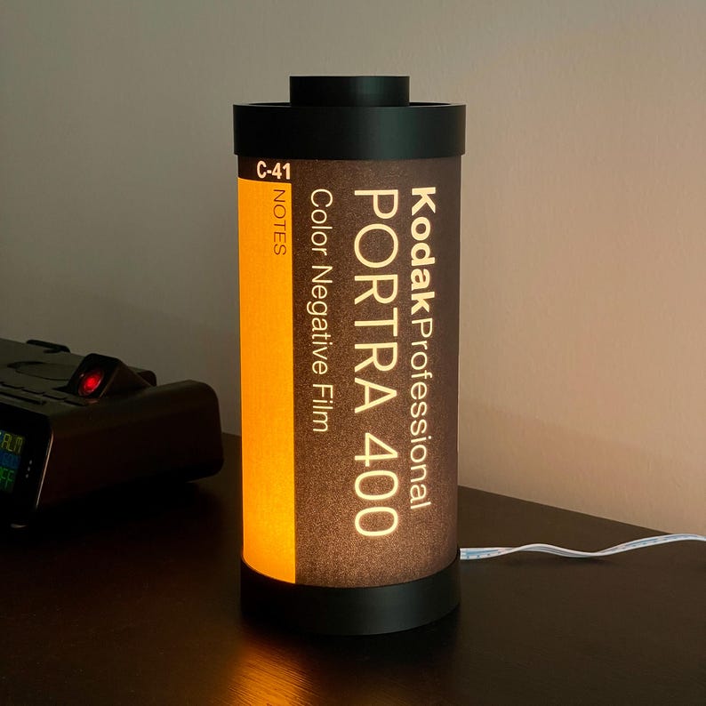 May include: A cylindrical lamp designed to resemble a Kodak Professional Portra 400 film canister. The lamp features a black top and base, with the film details printed on a brown and orange background. A white cord extends from the base.
