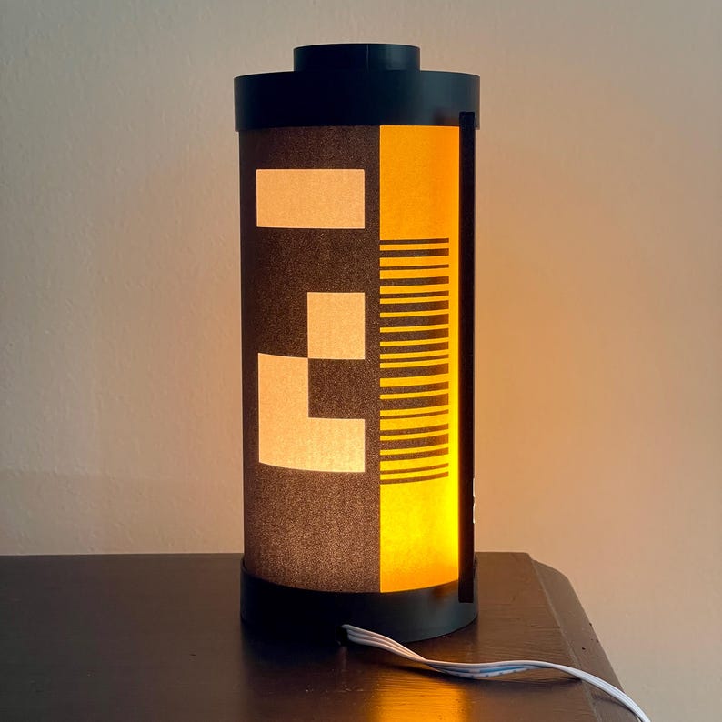May include: A cylindrical lamp with a black base and top, featuring a design reminiscent of a film canister. The lamp has a dark gray body with white geometric shapes and a yellow panel with black lines. A white cord extends from the base.
