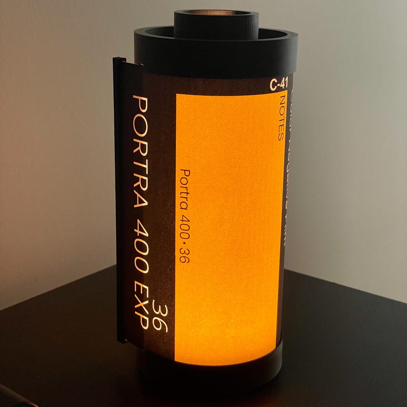 May include: A black lamp shaped like a 35mm film canister. The lamp has a bright orange center and features the text "PORTRA 400 EXP" and "36". The lamp is a unique home decor item.