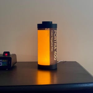 May include: A cylindrical lamp designed to resemble a roll of Portra 400 color negative film. The lamp has a black top and base, with the film canister label visible. The lamp emits a warm, orange glow.