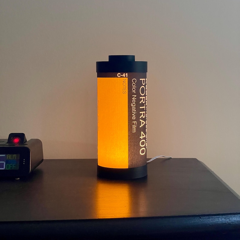 May include: A cylindrical lamp designed to resemble a roll of Portra 400 color negative film. The lamp has a black top and base, with the film canister label visible. The lamp emits a warm, orange glow.