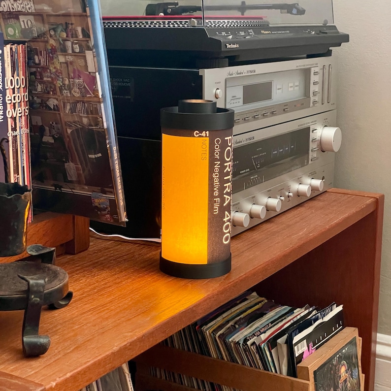 May include: A cylindrical lamp made from a film canister, with the text "PORTRA 400 Color Negative Film" and "C-41 NOTES". The lamp emits a warm, orange glow. It sits on a wooden shelf with vinyl records and audio equipment in the background.