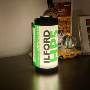 Könnte beinhalten: Eine zylindrische Lampe in Form einer Filmrolle mit dem Text "ILFORD HP5" in Grün auf weißem Hintergrund. Die Lampe hat einen schwarzen Deckel und Boden sowie ein grünes Etikett mit Filmangaben. Im Hintergrund eine New Yorker Taxi-Figur.