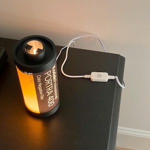 May include: A cylindrical lamp designed to resemble a Kodak Portra 400 film canister. The lamp has a black top and base, with the film details printed on the side. The lamp is lit from within, emitting a warm, orange glow. A white power cord with an on/off switch is attached.