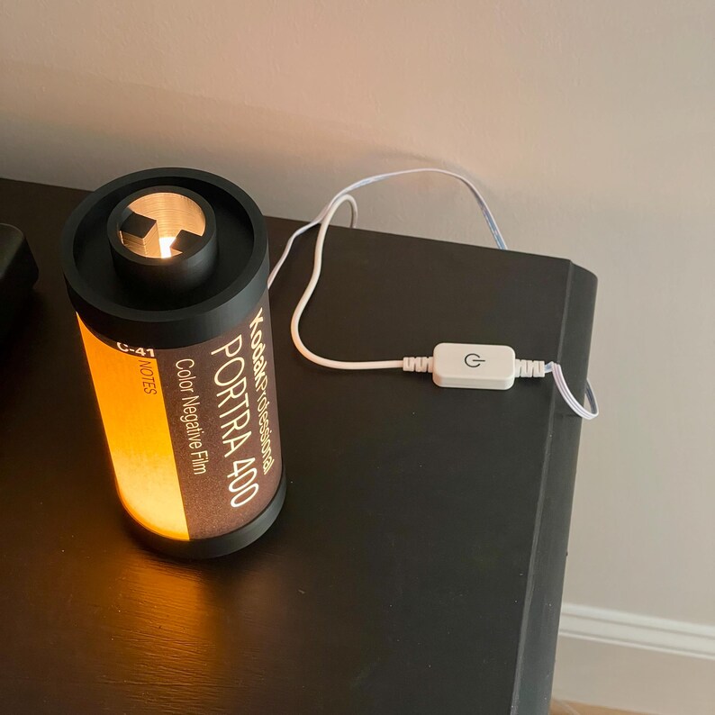 May include: A cylindrical lamp designed to resemble a Kodak Portra 400 film canister. The lamp has a black top and base, with the film details printed on the side. The lamp is lit from within, emitting a warm, orange glow. A white power cord with an on/off switch is attached.