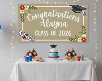 May include: A graduation party scene with a banner that reads "Congratulations Alayna CLASS OF 2026." The table is set with a cake, snacks, and red and blue cups. Balloons in red, white, and blue are on either side of the table.