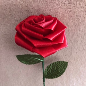 Puede incluir: Una rosa roja hecha a mano con cinta de raso, con hojas verdes y un tallo de alambre verde. La rosa está en plena floración, mostrando los intrincados pliegues de la cinta. Las hojas tienen vetas detalladas.