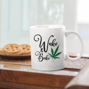 Puede incluir: Taza de café de cerámica blanca con texto negro que dice "Wake & Bake" y un gráfico de hoja de marihuana verde.