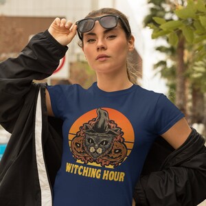 May include: A navy blue t-shirt with a retro-style graphic of a black cat wearing a witch's hat and surrounded by pumpkins. The text "Witching Hour" is printed below the graphic.
