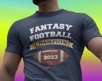 Op de afbeelding: Een donkerblauw t-shirt met de tekst "Fantasy Football Champion 2023" en een bruine voetbalafbeelding.