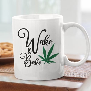 May include: White ceramic mug with the words "Wake & Bake" in black script, accompanied by a green cannabis leaf graphic. The mug has a curved handle and is set on a wooden surface.
