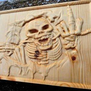 Può includere: Opera d'arte da parete in legno intagliato raffigurante uno scheletro con un teschio, una gabbia toracica e una mano tesa. L'opera è realizzata in legno chiaro e ha un bordo incorniciato. Lo scheletro è in un gesto.