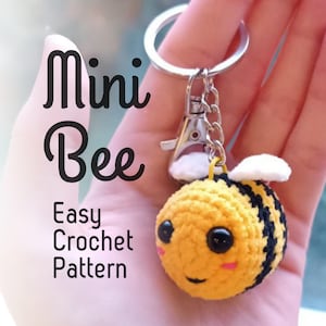Può includere: Un portachiavi a forma di ape all'uncinetto giallo e nero, con un viso sorridente, occhi neri e ali bianche. Il portachiavi ha un moschettone e un anello argentati. Il testo "Mini Bee Easy Crochet Pattern" è incluso.