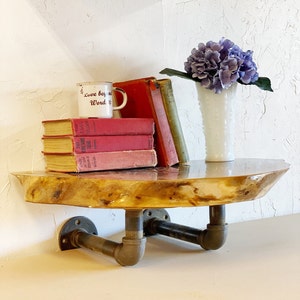 May include: A wooden shelf with a live edge, supported by black metal pipes. The shelf is decorated with a white mug with the text "Love beyond words", a stack of red books, and a white vase with purple flowers.