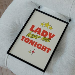 Peut inclure: Une affiche encadrée avec le texte "LADY hear me TONIGHT" en lettres rouges et jaunes, avec des étoiles et des croix. L'affiche est présentée sur un fauteuil blanc capitonné. L'arrière-plan comprend un mur bleu clair et un sol en bois.