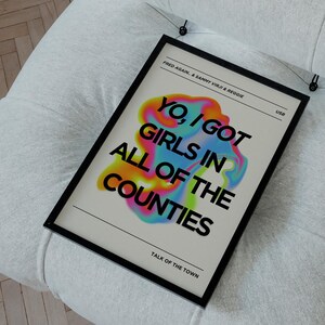 May include: A framed poster with the text "YO, I GOT GIRLS IN ALL OF THE COUNTIES" in colorful, stylized lettering. The poster is black-framed and hangs on a white, tufted chair. The text "TALK OF THE TOWN" is at the bottom.