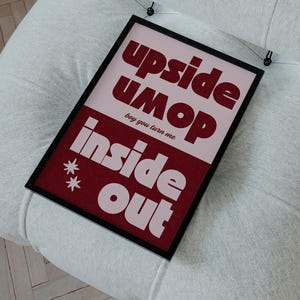 May include: A framed poster with the words "upside umop inside out" in red and white text, hanging on a white upholstered chair. The poster has a black frame and is suspended by two black clips.
