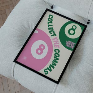 Puede incluir: Un póster enmarcado con el texto "COLLECT THE COMMAS" y un gráfico rosa y verde del número 8, apoyado en una silla tapizada blanca. El póster tiene un marco negro y está colgado de la silla.