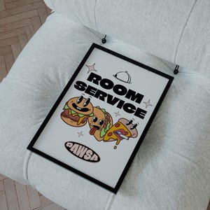 May include: A framed "Room Service" sign featuring cartoon burgers and pizza, hanging on a white upholstered chair. The sign has a black frame and the text "ROOM SERVICE" with a chef's hat icon. The sign also includes the text "AWSa".
