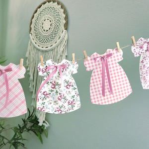 May include: A string of paper dresses in various patterns, including pink plaid, floral, and gingham, hang from a string with wooden clothespins. Each dress has a pink ribbon bow. A dreamcatcher and greenery are in the background.
