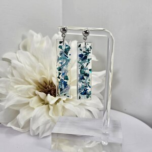 Op de afbeelding: Een paar zilveren oorhangers met helder hars en blauwe en zilveren glitter. De oorbellen worden getoond op een heldere acrylstandaard.