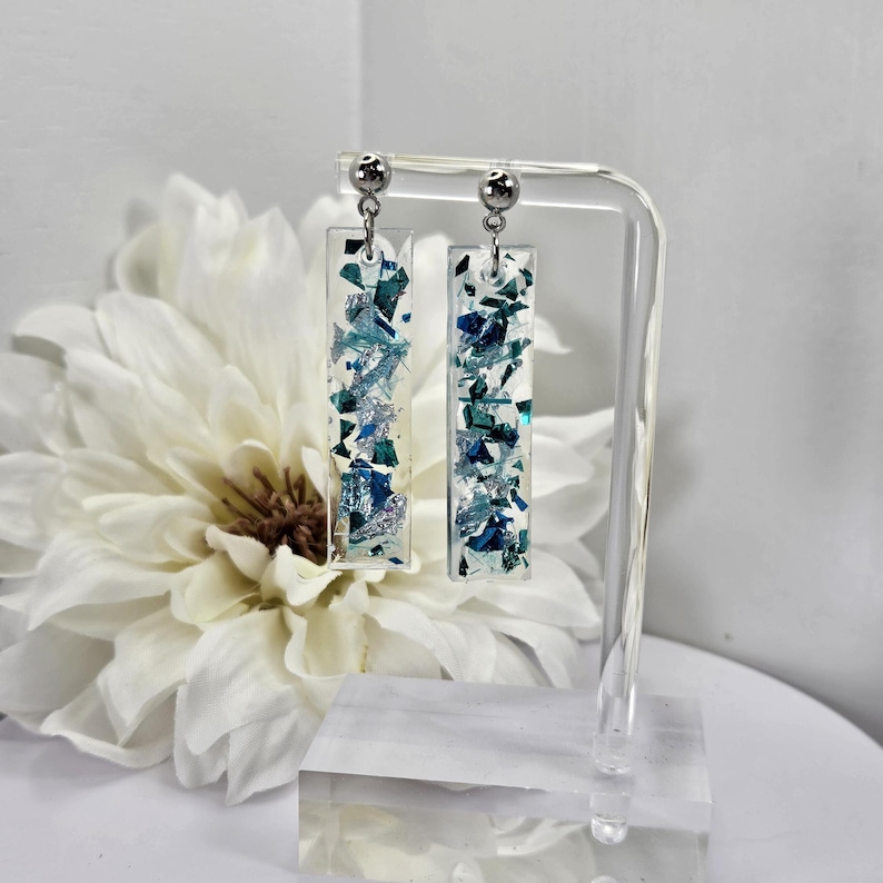 Op de afbeelding: Een paar zilveren oorhangers met helder hars en blauwe en zilveren glitter. De oorbellen worden getoond op een heldere acrylstandaard.