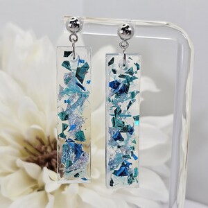 Op de afbeelding: Een paar zilveren oorhangers met helder rechthoekig hars dat blauwe en zilveren glitter bevat.