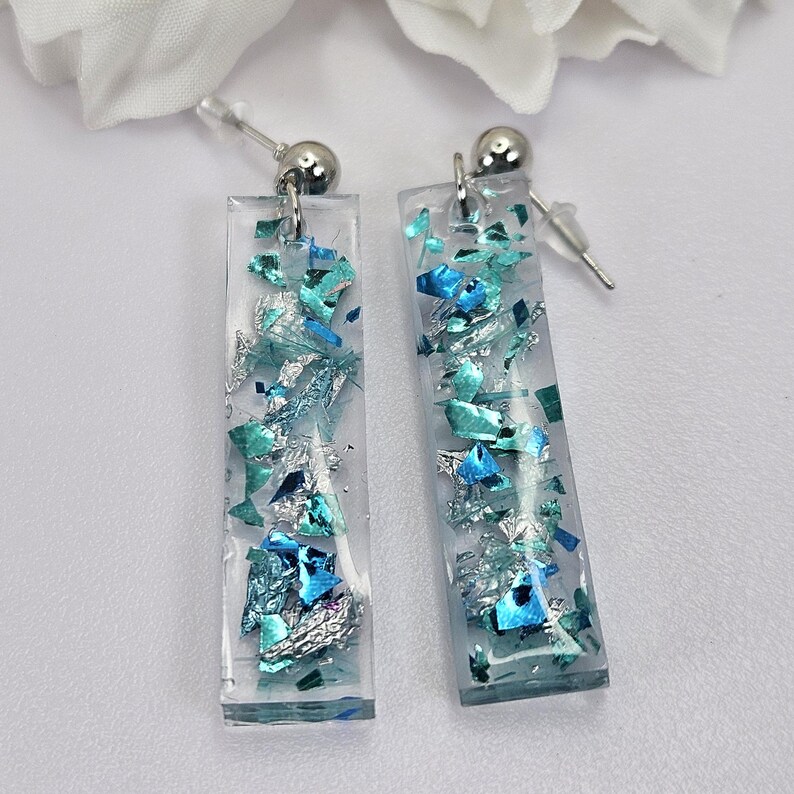 Op de afbeelding: Een paar zilveren oorhangers met rechthoekige, transparante harsbedels gevuld met blauwe en zilveren glitter.