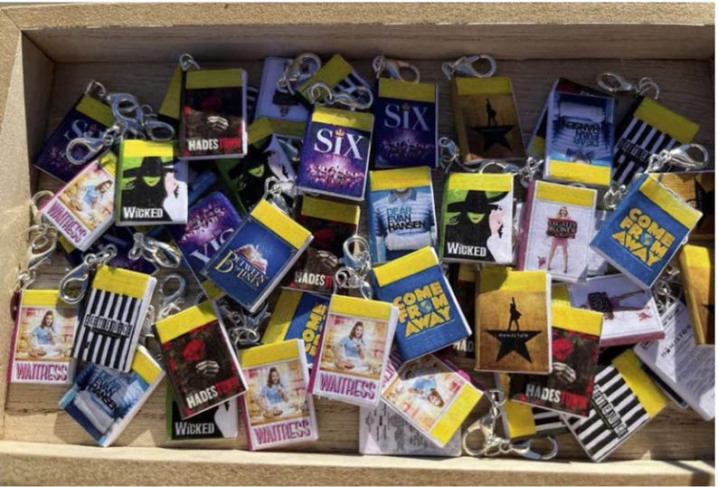 May include: A collection of miniature book-shaped keychains featuring various Broadway musical titles. The keychains have yellow and black striped edges, with images and text from shows like "Wicked," "Hamilton," and "Waitress."