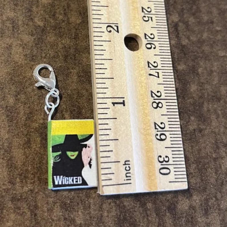 May include: Miniature charm of a book with the title "Wicked" on the cover, featuring a green-skinned witch and a woman in a pink dress. The charm is attached to a silver-colored lobster clasp. The book measures approximately 0.75 inches in height.
