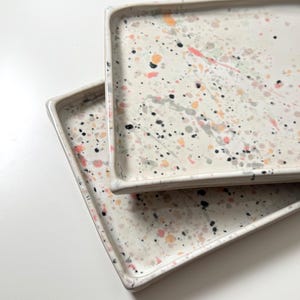 Könnte beinhalten: Zwei rechteckige Tabletts mit gesprenkeltem Design. Die Tabletts sind weiß mit einem Muster aus schwarzen, grauen, rosa und orangefarbenen Farbspritzern. Die Tabletts haben einen erhöhten Rand.