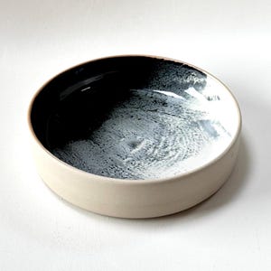 Könnte beinhalten: Eine runde Keramikschale mit cremefarbenem Äußeren und einem schwarz-weiß-grauen Innendesign. Das Innere der Schale weist ein strukturiertes, abstraktes Muster auf, das einen auffälligen Kontrast zum glatten, neutralen Äußeren bildet.