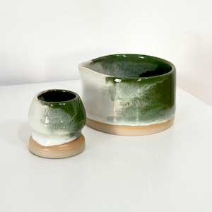 Könnte beinhalten: Eine kleine Keramiktasse und eine größere Keramikschale mit Ausguss. Beide Stücke haben eine glänzende weiße und waldgrüne Glasur mit einem verwaschenen, gesprenkelten Finish und einen unglasierten, glatten beigen Boden an der Unterseite.