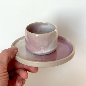 Könnte beinhalten: Ein handgefertigtes Keramik-Tassen- und Untertassen-Set. Die Tasse ist zylindrisch mit einer hellvioletten Glasur, die am Rand in Weiß übergeht. Die Untertasse hat eine violette Mitte und einen hellbeigen Rand. Das Set wird von einer Hand gehalten.