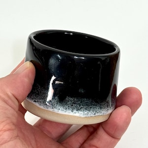 Könnte beinhalten: Ein kleiner Keramikbecher mit einer glänzend schwarzen Glasur im oberen Bereich, die in ein strukturiertes, weiß gesprenkeltes Band nahe der Basis übergeht, welche in einem natürlichen, unglasierten beigen Ton mit glattem Rand gehalten ist.