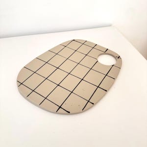 Könnte beinhalten: Ein glattes, beigefarbenes, oval geformtes Keramikbrett mit einem handgemalten schwarzen Gittermuster auf der Oberfläche. Das Brett verfügt über eine einzelne kreisförmige Aussparung nahe dem rechten Rand, die zum Aufhängen oder Greifen dient.