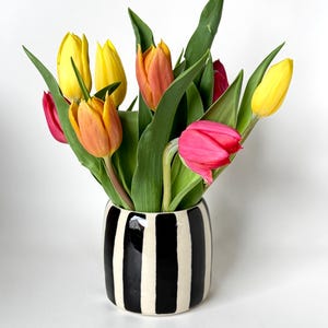 Könnte beinhalten: Ein bunter Strauß Tulpen in einer schwarz-weiß gestreiften Vase. Die Tulpen zeigen Farben wie Gelb, Orange und Rosa, mit grünen Blättern. Die Vase ist zylindrisch und hat vertikale Streifen.