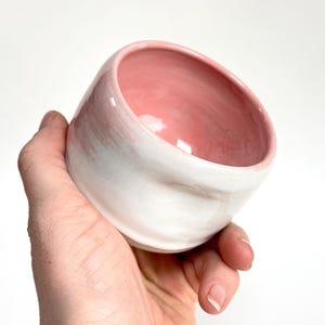 Könnte beinhalten: Handgefertigte Keramikschale mit weißer Außenseite und rosa Innenseite. Die Schale hat eine abgerundete, asymmetrische Form. Die Schale wird in einer Hand vor weißem Hintergrund gehalten.