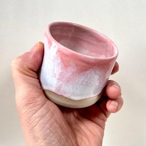 Könnte beinhalten: Eine handgefertigte Keramiktasse mit rosa und weißer Glasur. Die Tasse hat einen natürlichen, unglasierten Boden und wird in einer Hand gehalten. Der Innenraum ist zartrosa, wobei die Farbe zum Rand hin in Weiß übergeht. Die Tasse ist etwa 7.6 cm hoch.