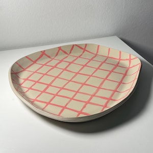 Könnte beinhalten: Ein quadratischer Keramikteller mit abgerundeten Ecken, einer cremefarbenen Basis und einem handgemalten Gittermuster in leuchtendem Pink. Die Oberfläche hat einen leicht unregelmäßigen, organischen Rand und ungleichmäßig verlaufende Linien.