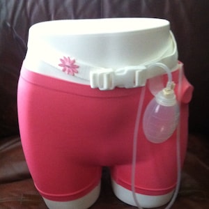 May include: Pink underwear with a white elastic waistband and a white plastic belt with a pink flower design. A clear plastic bag is attached to the belt with a clear plastic tube.
