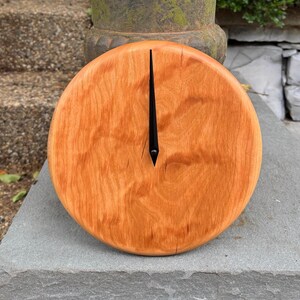May include: A round wooden clock with a natural wood grain finish. The clock face is a warm orange-brown color, and it has a single, long, black hand pointing upwards. The clock is displayed on a gray stone surface.