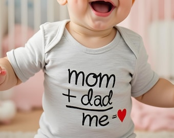 May include: A cheerful baby wearing a light grey onesie with the text "mom + dad = me" in black, with a red heart. The baby is in a nursery with a cot and pink decor.