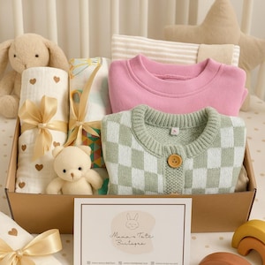 Peut inclure: Un coffret cadeau pour bébé contenant un sweat-shirt rose, un cardigan à carreaux vert et blanc, des couvertures d'emmaillotage, un ours en peluche et un lapin en peluche. La boîte comprend une carte avec le texte "Mama's Tate Boutique".