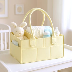 May include: A pale yellow felt storage caddy with two handles and multiple exterior pockets. The interior contains a cream-coloured plush rabbit toy, a stack of white nappies, a light blue bottle, and a white baby bottle with a clear teat.
