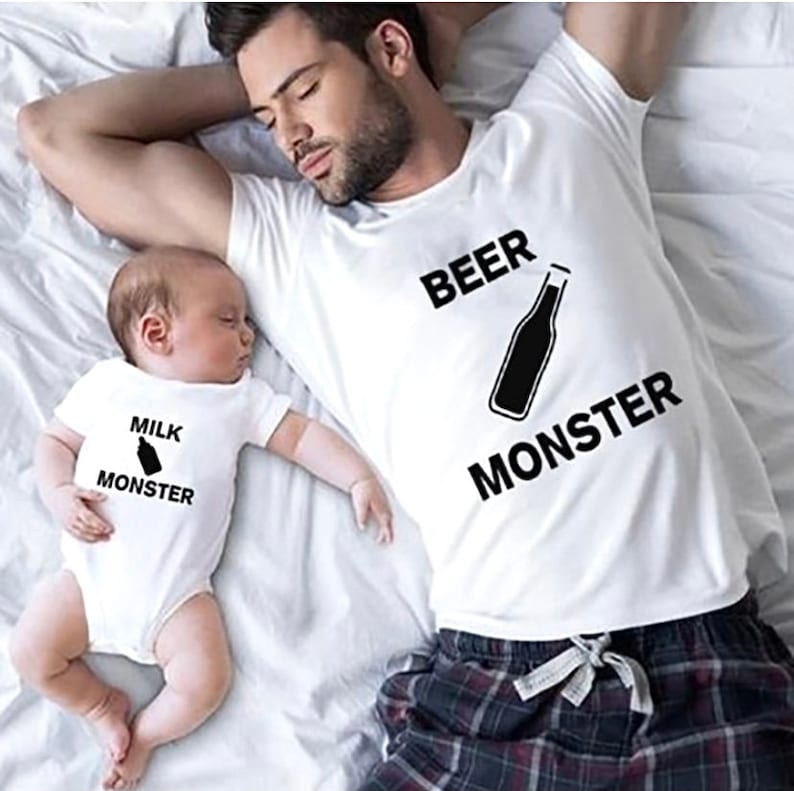 Op de afbeelding: Een slapende man en baby dragen bijpassende witte T-shirts. Op het shirt van de man staat "BEER MONSTER" met een flesafbeelding. Op het shirt van de baby staat "MILK MONSTER" met een flesafbeelding. De man draagt een geruite pyjamabroek.