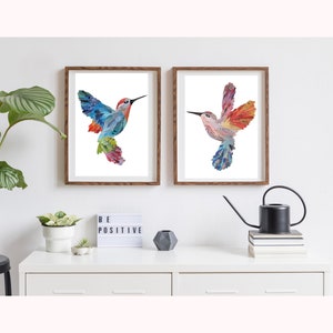 May include: Two framed prints of colorful hummingbird illustrations. The hummingbirds are made of paper and have a layered, textured look. The prints are in brown frames and are hanging on a white wall.